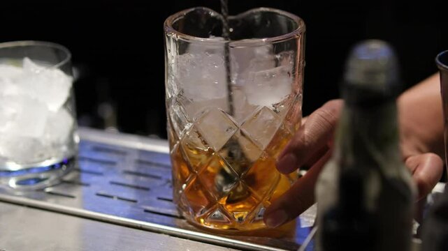 Close up of a professional bartender is mixing an alcoholic cocktail with professional crystal long lasting ice cubes to customers at the bar or disco club. Professional bartender masterly creating