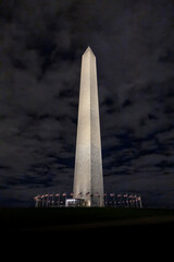 The Washington Monument illuminated against a dark, cloudy night sky, surrounded by American flags.