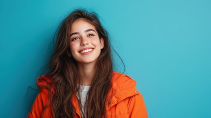 Fototapeta premium Happy young Latino woman with long brown hair wearing a bright orange jacket standing against a solid blue background