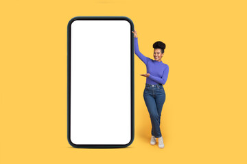 A young Brazilian woman in casual attire stands beside a large blank phone, smiling and gesturing. The vibrant yellow background highlights her youthful energy and style.