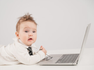 A cute child wearing a suit holds glasses and lies behind a laptop.