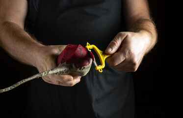 A pair of focused hands carefully peels a rich red beetroot using a bright yellow peeler. The backdrop is dark, highlighting the vivid colors and the intricate motions of preparing fresh ingredients