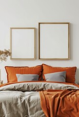 A minimalistic bedroom with a bed. two empty frames. and a vase with dried flowers The bed has a gray duvet and two orange pillows On the bed is an orange blanket