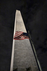 Washington Monument at night with the American flag waving prominently against a cloudy sky, showcasing national pride and historical significance.