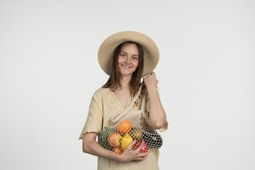 A cheerful woman wearing a straw hat stands in a minimalist studio, holding a net bag filled with various vibrant fruits, radiating joy and health