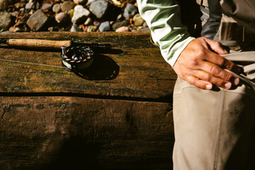 Closeup of a fly fishing rod next to the hand and leg of a fisherman sitting on a large tree trunk on the Pecos River, New Mexico