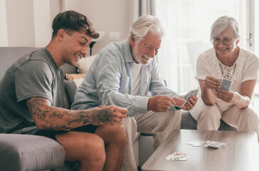 Happy multigenerational family of white-haired grandparents and young grandson play cards sitting...