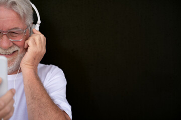 Portrait of half senior bearded man smiling while listening to music by smart phone.  White headphones, black background. Copy space for text