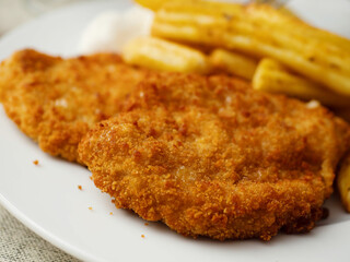 Breaded chicken fillet meal and fried potatoes chips with ridges on a white plate and light color table cloth. Simple food made of high quality premium products. Cooked poultry meat.