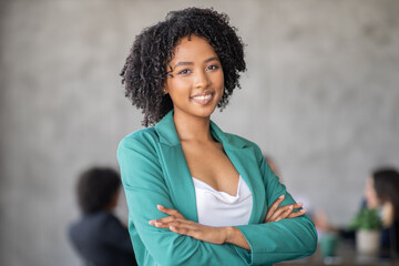 Fototapeta premium A confident woman in a green blazer stands with her arms crossed, showcasing teamwork in a modern office. A diverse group engages in discussion, emphasizing cooperation and success.