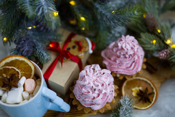 Pink cakes on a wooden board under a Christmas tree with lights. A cup with marshmallows and orange slices
