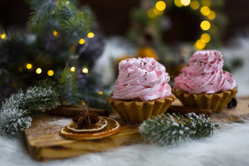 Pink cakes on a wooden board under a Christmas tree with lights. A cup with marshmallows and orange slices