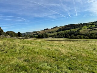 A lush green meadow stretches across rolling hills under a vibrant blue sky. Thin clouds streak the sky, as sunlight casts a warm glow over the countryside near, Goose Eye, UK