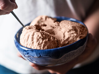 A close-up of a blue ceramic bowl filled with scoops of creamy ice cream, held in hands with a spoon