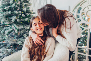 Happy mother kissing her teenager daughter at home by decorated Christmas tree. Family celebrating holiday