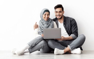 Loving muslim family sitting on floor in new apartment with laptop, buying furniture for house or working on design together, sitting by blank wall, hugging and looking at computer screen, free space