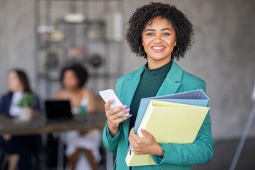A confident businesswoman stands in a modern office, holding folders and a smartphone. Behind her,...