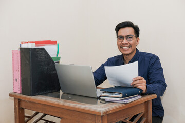 Smiling Southeast Asian Man Working While Sitting In Front Of Laptop In Home Office with hand holding paperwork. Workplace Of Professional Worker, Freelancer or Student.