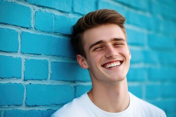 A smiling young man leans against a blue brick wall, looking relaxed and friendly