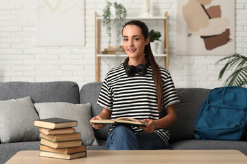 Beautiful happy female student with stack of books, backpack and headphones sitting on sofa at home