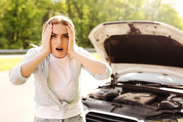 Emotional young blonde woman standing near broken car with open hood, grabbing her head in terror,...