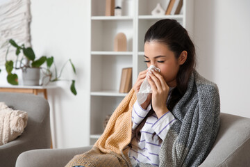 Ill young woman with tissue sneezing in armchair at home