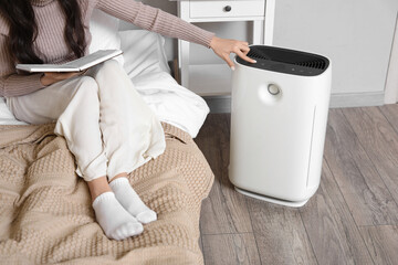 Young woman with book turning on air purifier in bedroom