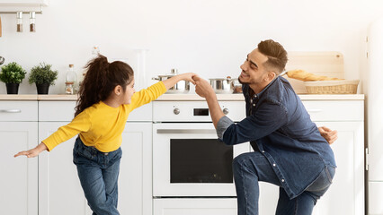 Cheerful arab father dancing with his cute little daughter at home, happy middle eastern dad having fun and fooling with child in kitchen interior, standing one one knee and smiling, copy space