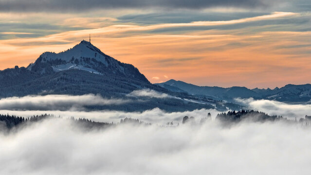 Drohnenaufnahme im Winter mit Nebel im Tal und Sonne im Gebirge im Sonnenuntergang
