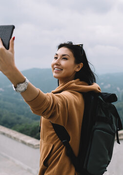 Smiling asian female tourist in casual wear and backpack making picture on mobile phone camera during vacations, cheerful woman posing for selfie exploring destinations in journey chare content online