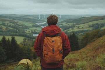 Naklejka premium A man in a red jacket and orange backpack stands on a hillside, gazing at a sprawling green valley with rolling hills, trees, and distant wind turbines.