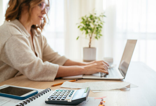 Beautiful middle-aged woman in glasses making marks in financial documents at home office using laptop. Small business, home finances, money savings concept image.