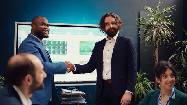 Business partners celebrate the beginning of a new project between their companies, shaking hands in a meeting. Leaders forging an agreement to develop the corporation and collect resources. Camera B.