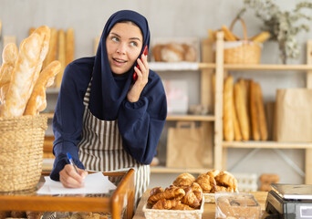 Young woman with European features, wearing blue hijab and striped apron, working in small bakery filled with variety of baked goods, speaking on phone while managing orders