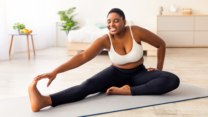 Fototapeta premium Curvy black lady stretching her legs, exercising on yoga mat, leading active lifestyle, trying to lose weight at home. Plus size young African American woman working out during her slimming program