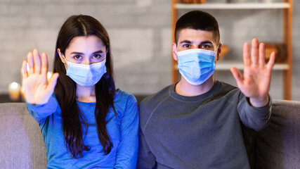 Stop Coronavirus Outbreak. Young couple in protective surgical masks gesturing stop sitting on the couch at home with pc