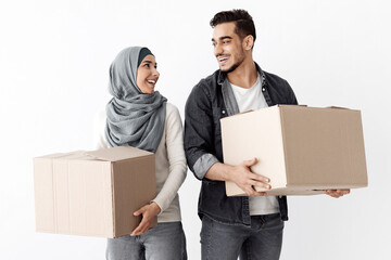 Happy young arab husband and wife in hijab holding paper boxes with belongings, moving to new apartment, looking at each other and cheerfully smiling, white background. Relocation, new house caoncept