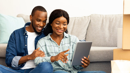 Cheerful Black Spouses With Digital Tablet Sitting Among Cardboard Boxes After Moving To New Flat, Smiling African American Couple Using Tab Computer For Online Shopping Or Browsing Internet