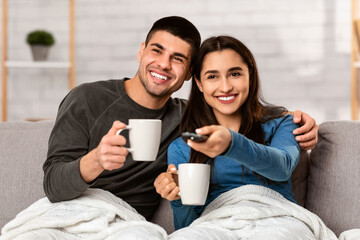 Happy couple drinking hot tea or coffee and watching TV at home, wrapped in warm blanket, copy space