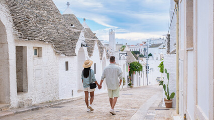 A couple walks hand in hand along a narrow cobblestone street lined with whitewashed trulli houses,...