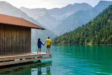 A diverse couple stands hand in hand on a wooden dock, surrounded by majestic mountains and crystal-clear water. The serene atmosphere of Plansee Austria invites peaceful reflection and connection.