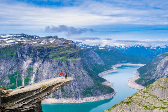 Two individuals stand on a rock outcrop overlooking a valley with a winding lake, surrounded by snow-capped mountains. Trolltunga, Norway, A diverse couple of men and women on vacation
