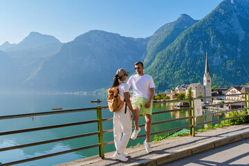 A diverse couple of men and women shares a light moment by the tranquil lake in Hallstatt, Austria, surrounded by majestic mountains and charming architecture under a bright blue sky.