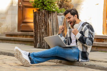 Handsome Hispanic young man using laptop computer celebrating win good message news, lottery jackpot victory, giveaway online outdoors. Happy Indian guy tourist sitting on city street. Town lifestyles