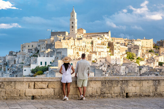 A couple stands on a stone balcony, gazing at the breathtaking skyline of Matera Italy. The ancient buildings bask in the warm glow of sunset, a diverse couple of men and woman on a city trip - Powered by Adobe