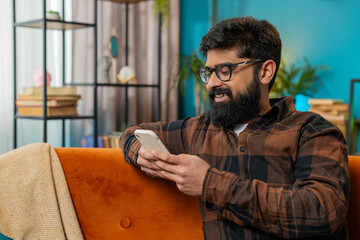 Happy young man in casual clothes thinking, text messaging on smartphone. Indian guy sitting on sofa and using cellphone in living room at home. Male guy is searching information on browser internet