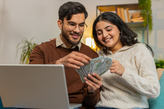 Happy young Hispanic couple counting money and booking tickets for holiday vacation sits on sofa in living room at home. Diverse family holding banknotes planning online investment budget home finance