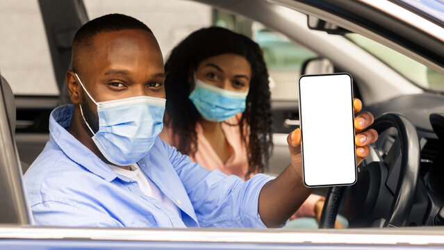Closeup of black man in surgical mask showing cell phone with white empty screen for mock up sitting on driver seat in car with wife, holding gadget in hand, copy space template. Health passport - Powered by Adobe