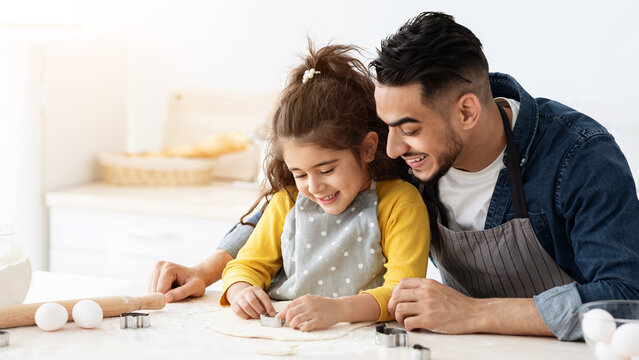 Cute little arab girl cutting out different cookie shapes from dough while baking with dad in kitchen, happy middle eastern father and daughter enjoying cooking pastry at home together, copy space