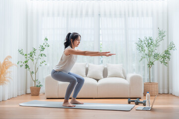 Young Asian beautiful woman doing aerobic exercise at home. Attractive girl doing  workout by squatting, follow instructions video from online trainer for health in house